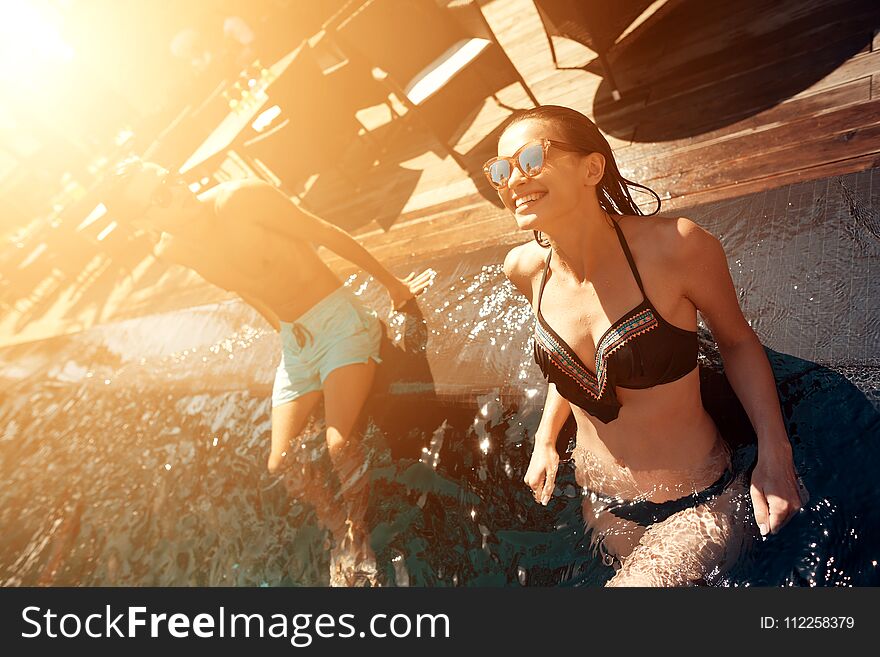 Young happy couple is resting under sun in swimming pool at summer time. Company of friends carefree spend time swimming in pool.