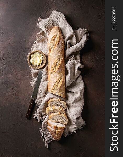 Loaf of sliced fresh baked artisan baguette bread on linen cloth with butter, herbs, pink salt and vintage knife over dark brown texture background. Top view, copy space.