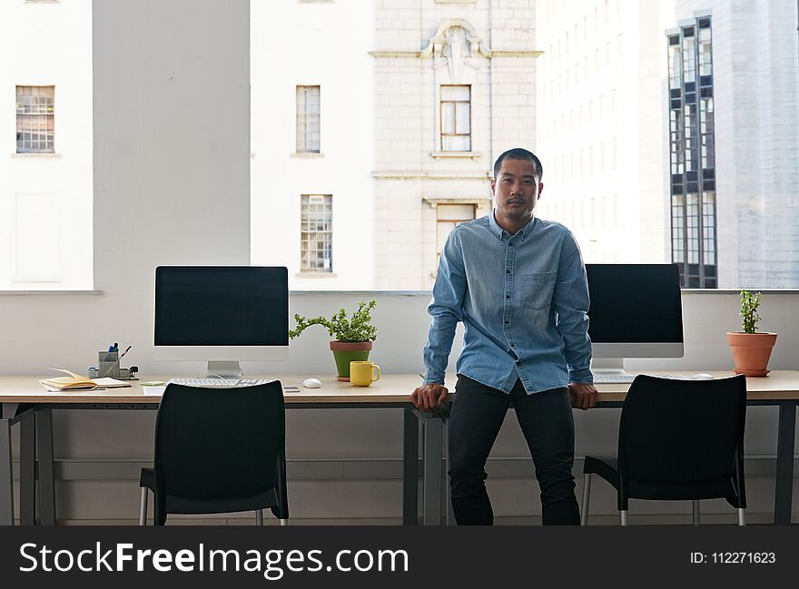 Content young Asian businessman leaning on a table by computers while working alone in a modern office. Content young Asian businessman leaning on a table by computers while working alone in a modern office