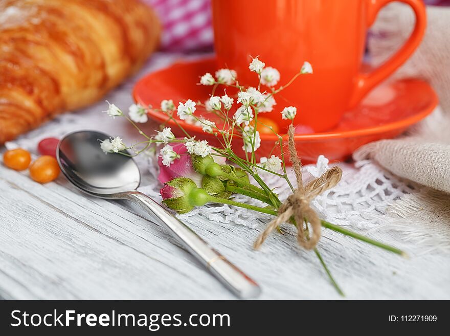 Cup of coffee and croissant are decorated by napkins, roses and candies on a white wooden table. Romantic atmosphere. Cup of coffee and croissant are decorated by napkins, roses and candies on a white wooden table. Romantic atmosphere