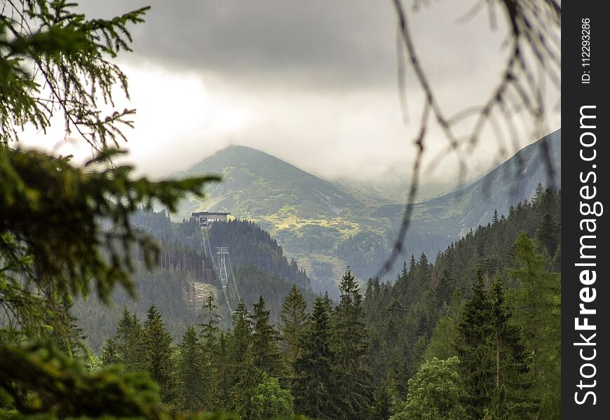View from route to Nosal peak in Tatras. Forest and trees in the foreground and some peaks of Tatra range in the background. Cable way to Kasprowy Wierch mountain in Poland. Dark cloudy sky and fog . Photo taken during rainy summer day. View from route to Nosal peak in Tatras. Forest and trees in the foreground and some peaks of Tatra range in the background. Cable way to Kasprowy Wierch mountain in Poland. Dark cloudy sky and fog . Photo taken during rainy summer day.