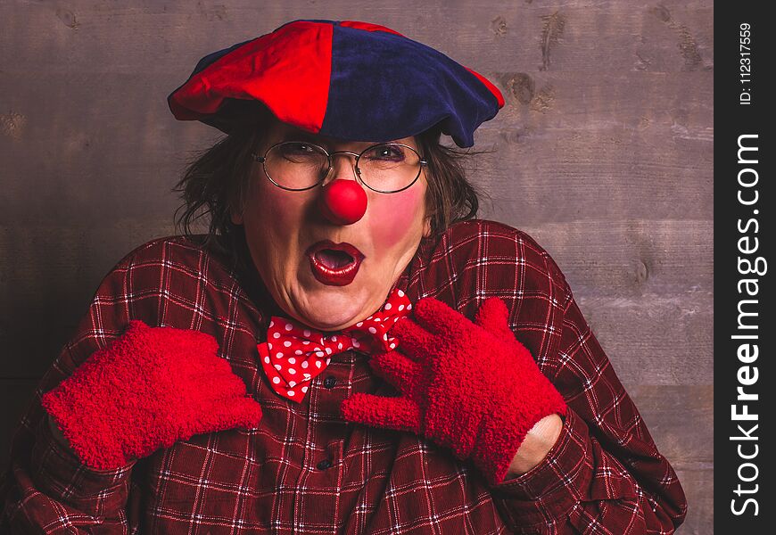 Woman emotional funny cheerful clown with glasses, red nose, shirt, hat and gloves.Wooden background. Woman emotional funny cheerful clown with glasses, red nose, shirt, hat and gloves.Wooden background