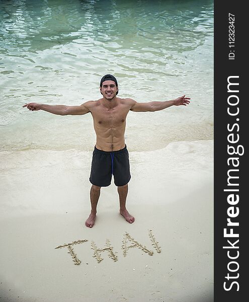 Joyful young man standing near thai letters on a sandy shore. Vertical outdoors shot. Joyful young man standing near thai letters on a sandy shore. Vertical outdoors shot.