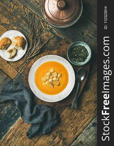 Flat-lay of fall warming pumpkin cream soup with croutons and seeds on board over rustic wooden background, top view, vertical composition. Autumn vegetarian, vegan, healthy comfort food concept. Flat-lay of fall warming pumpkin cream soup with croutons and seeds on board over rustic wooden background, top view, vertical composition. Autumn vegetarian, vegan, healthy comfort food concept