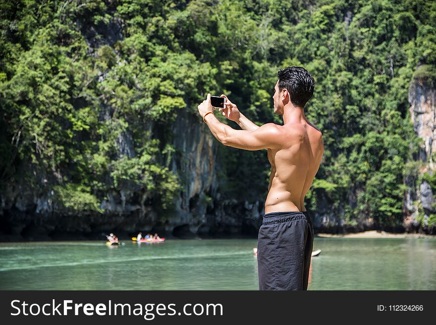 Half body shot of a handsome young man using cell phone, standing on a beach in Phuket Island, Thailand, shirtless wearing boxer shorts, showing muscular fit body. Half body shot of a handsome young man using cell phone, standing on a beach in Phuket Island, Thailand, shirtless wearing boxer shorts, showing muscular fit body