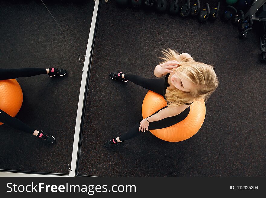 Seductive blonde woman, dressed in dark fitness clothing, posing on large orange exercise ball in front of the mirror