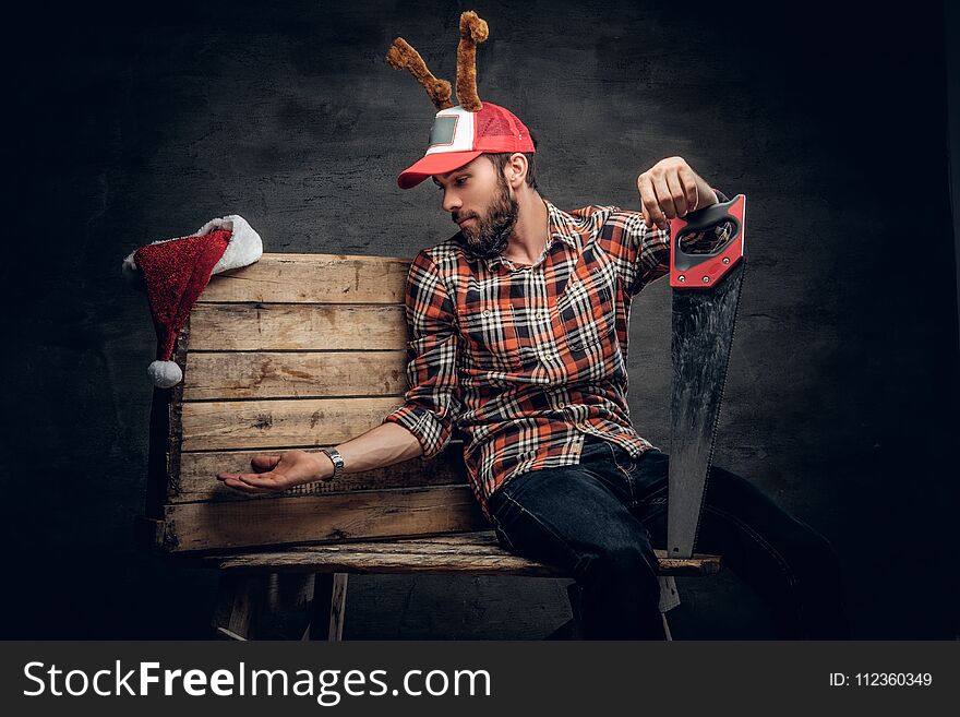 A bearded male in Christmas cap with deer horns posing on wooden palette and holds handsaw. A bearded male in Christmas cap with deer horns posing on wooden palette and holds handsaw.