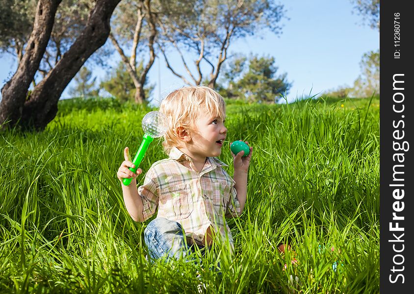 Funny toddler boy pretends to bite a colored Easter egg in the green grass. Funny toddler boy pretends to bite a colored Easter egg in the green grass