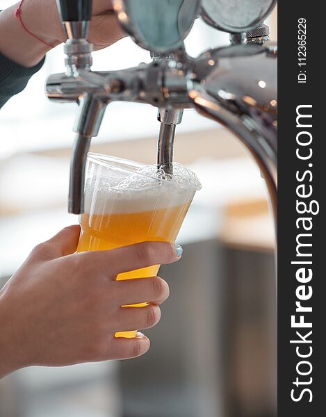 Plastic cup of beer flowing from beer tap with nice foam close-up in female bartender hands on blurred background