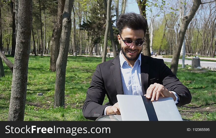 Attractive person sits by table, smiling with dimples on cheeksand uses laptop. Guy has short dark hair, beard, dimples on cheeks and wears sunglasses. Concept of good price of massage modern technologies. Attractive person sits by table, smiling with dimples on cheeksand uses laptop. Guy has short dark hair, beard, dimples on cheeks and wears sunglasses. Concept of good price of massage modern technologies.