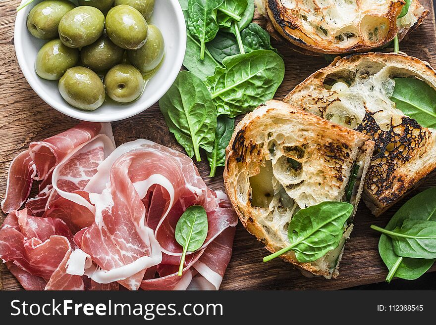 Close up prosciutto, olives, grilled mozzarella spinach sandwiches on wooden background, top view. Mediterranean style snack, appetizer, tapas