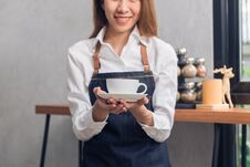 Close Up Of A Young Asian Female Barista Hold A Cup Of Coffee Serving To Her Customer With Smile Surrounded With Bar Counter. Royalty Free Stock Photos