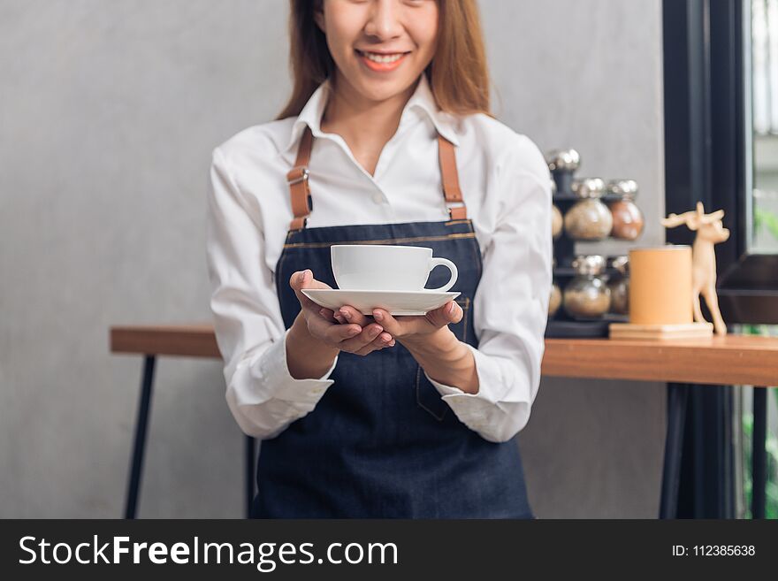 Close up of a young Asian female barista hold a cup of coffee serving to her customer with smile surrounded with bar counter.