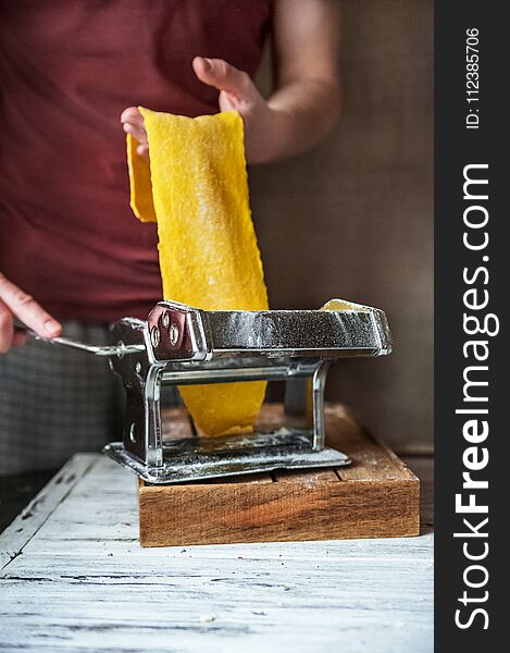 These multicolored pasta recipes are dyed with organic vegetables and spices on a wooden background. Preparation of yellow lasagna at home with parsley and basil, male hands cut pasta on a white wooden table