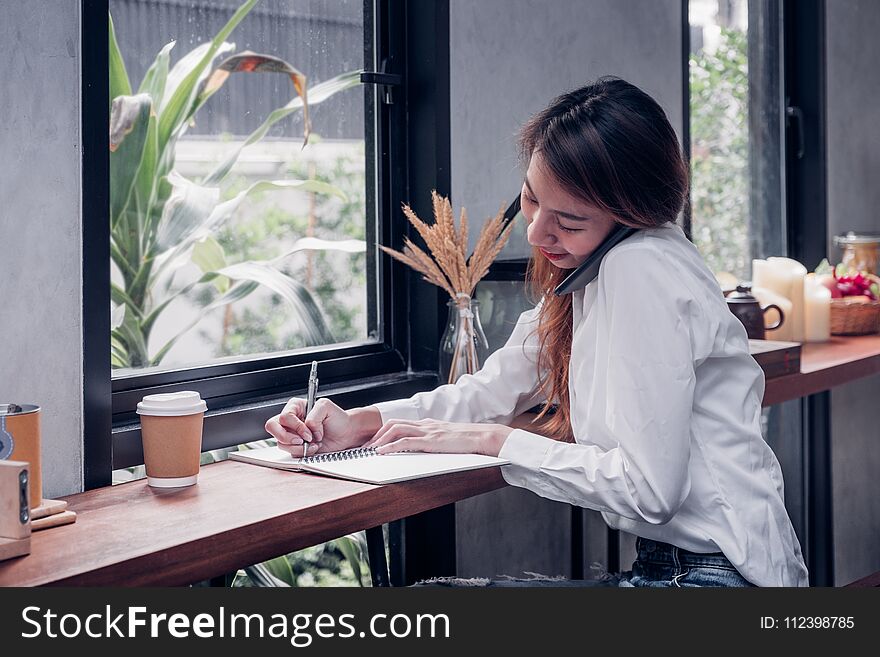 Asian Businesswoman use mobile phone contact customer and writing a note on wood table at coffee shop near window,woking outside