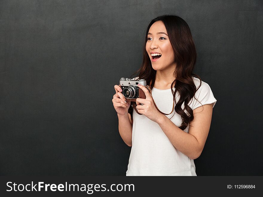 Image of asian young excited woman photographer holding camera isolated over grey wall background. Looking aside.