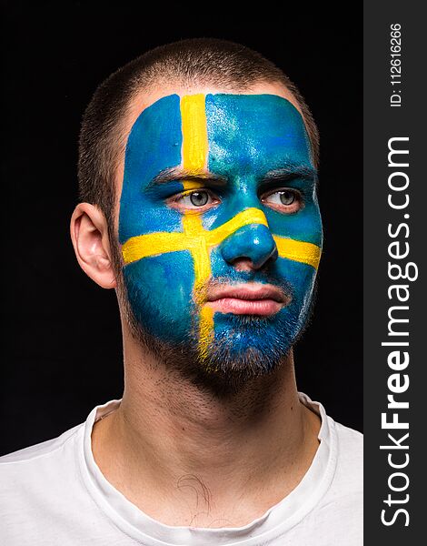 Portrait of handsome man face supporter fan of Sweden national team with painted flag face isolated on black background. Fans emot