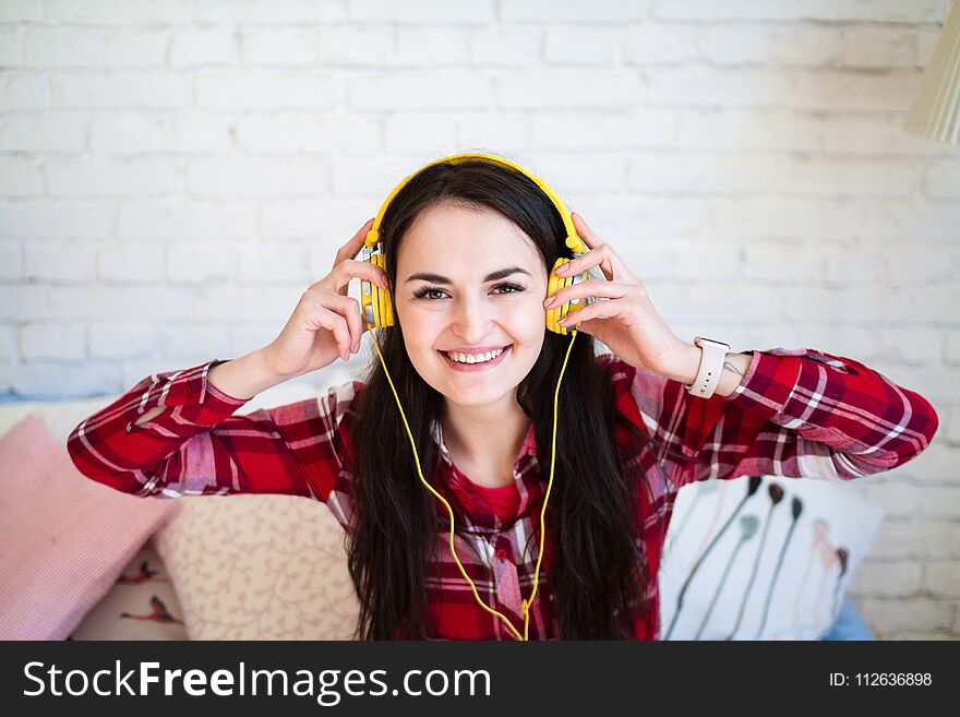 Portrait of a beautiful woman in the morning listening to music sitting on bed at home. Red plaid shirt, yellow headphones, white brick wall. Portrait of a beautiful woman in the morning listening to music sitting on bed at home. Red plaid shirt, yellow headphones, white brick wall.