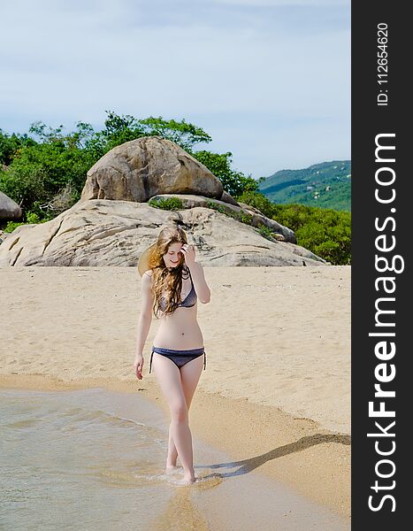 Blonde in a bikini on a paradise beach. The mountains are on the horizon. The sky is sun-drenched.