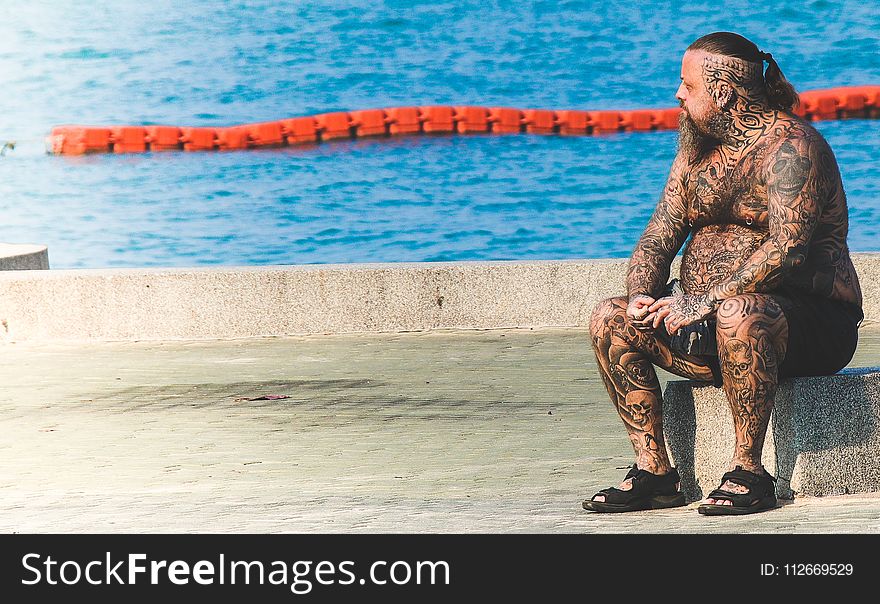 Man With Tattoos Sitting On Gray Concrete Floor Near Body Of Water