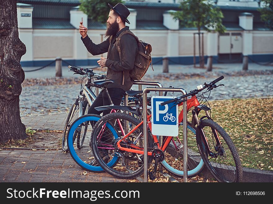 Handsome hipster traveler with a stylish beard and tattoo on his arms dressed in casual clothes and hat, using the phone, standing on the bicycle parking. Handsome hipster traveler with a stylish beard and tattoo on his arms dressed in casual clothes and hat, using the phone, standing on the bicycle parking.