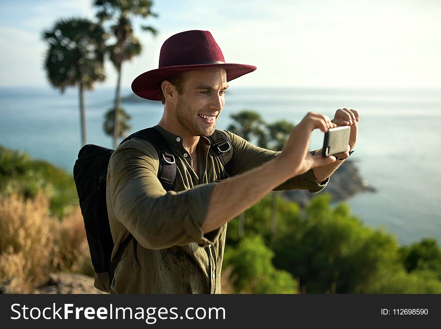 Happy man with a black backpack takes a photo on his cellphone on the background of the palms and the sea. He wears an olive shirt and crimson hat. Sun shines onto his body. Horizontal. Happy man with a black backpack takes a photo on his cellphone on the background of the palms and the sea. He wears an olive shirt and crimson hat. Sun shines onto his body. Horizontal.