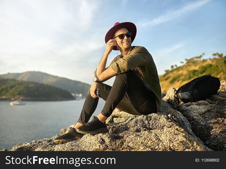 Nice smiling guy in sunglasses is listening music while sitting on the rocky cliff on the sunny background of the sea bay. He wears dark jeans and sneakers, olive shirt and crimson hat. Horizontal. Nice smiling guy in sunglasses is listening music while sitting on the rocky cliff on the sunny background of the sea bay. He wears dark jeans and sneakers, olive shirt and crimson hat. Horizontal.