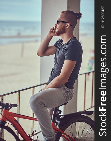 Handsome redhead male with a stylish haircut and beard dressed in sportswear with a bicycle, talking on the phone standing outside. Handsome redhead male with a stylish haircut and beard dressed in sportswear with a bicycle, talking on the phone standing outside.