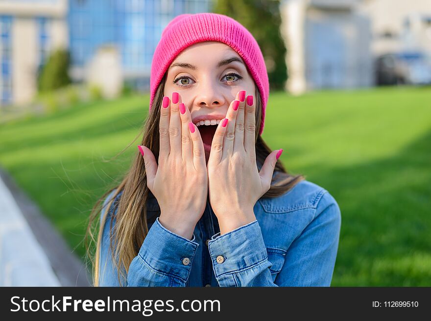 Happy joyful surprised teenage girl in pink hat, with pink nails, dressed in jeans shirt covering her mouth with palms