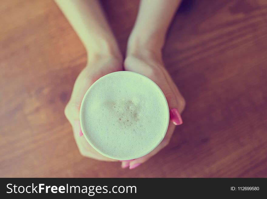 Woman`s hands holding a cup of frothy coffee over wooden background, top view