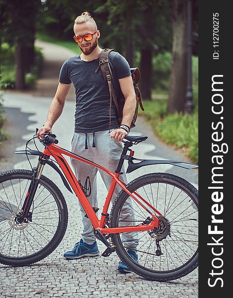 Handsome redhead male with a stylish haircut and beard dressed in sportswear and sunglasses walks in the park with a bicycle and backpack. Handsome redhead male with a stylish haircut and beard dressed in sportswear and sunglasses walks in the park with a bicycle and backpack.