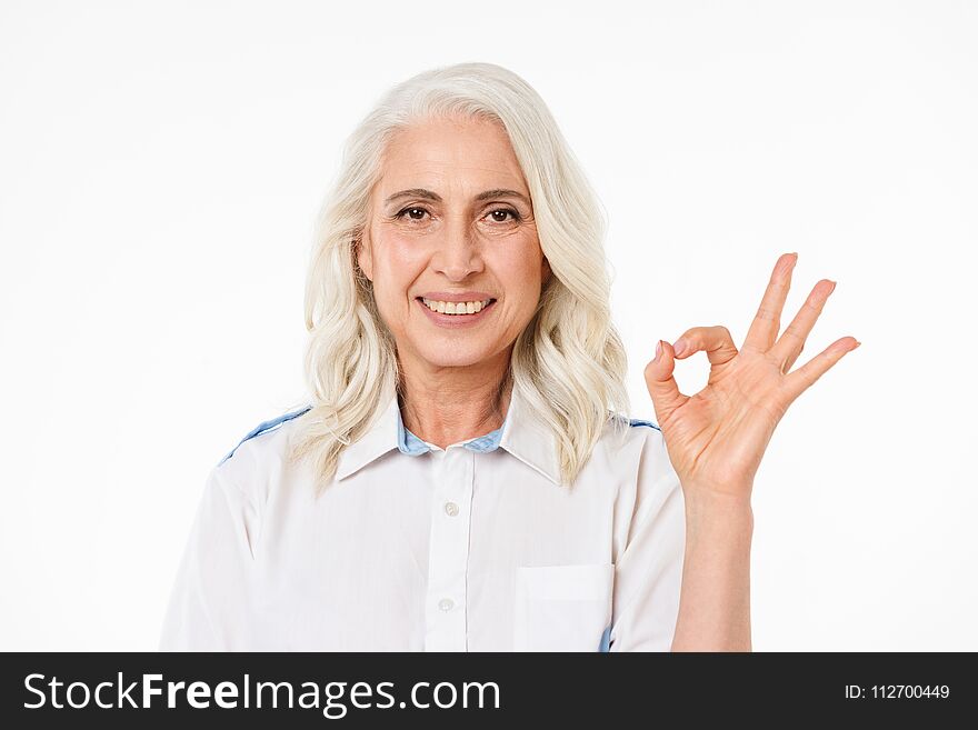 Image of mature old smiling woman standing isolated over white background looking camera showing okay gesture. Image of mature old smiling woman standing isolated over white background looking camera showing okay gesture.