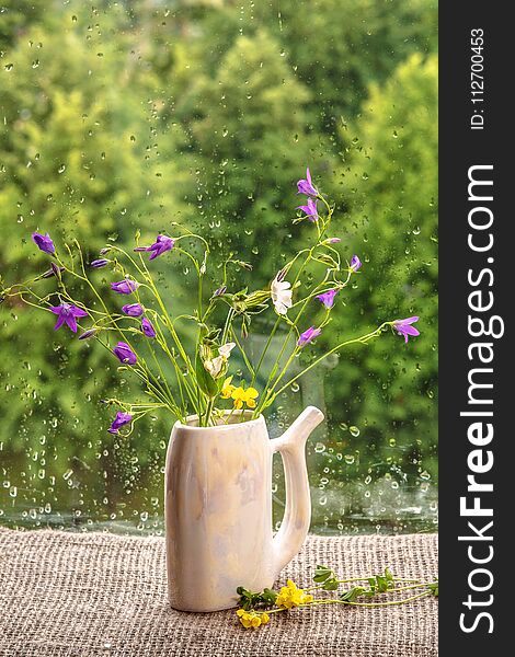 A bouquet of wild flowers stands on the window sill. On glass rain drops. Purple flowers bells on a natural green background. Rustic style. A bouquet of wild flowers stands on the window sill. On glass rain drops. Purple flowers bells on a natural green background. Rustic style.