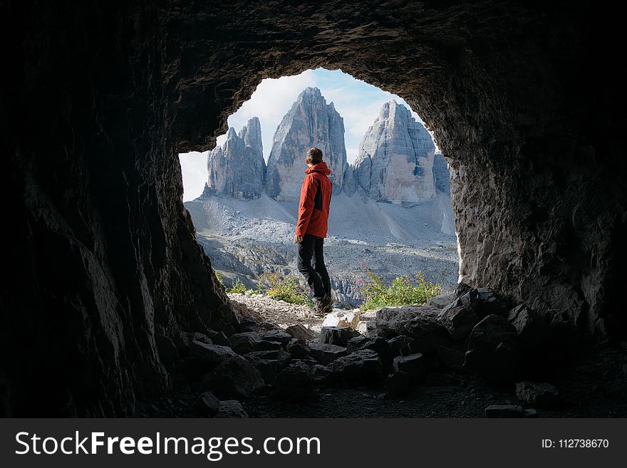 Man In Red Jacket Standing Outside Of Cave In Front Of Three Mountains