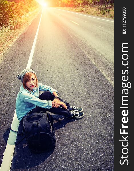 Rear view of a young woman hitchhiking carrying backpack sitting on the road