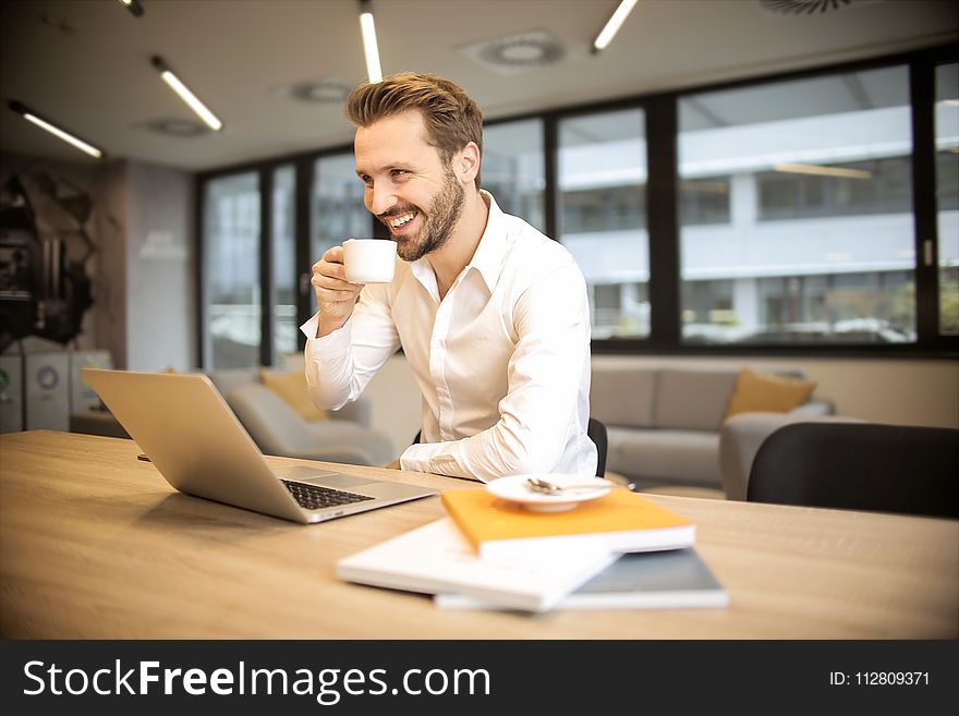Depth of Field Photo of Man Sitting on Chair While Holding Cup in Front of Table
