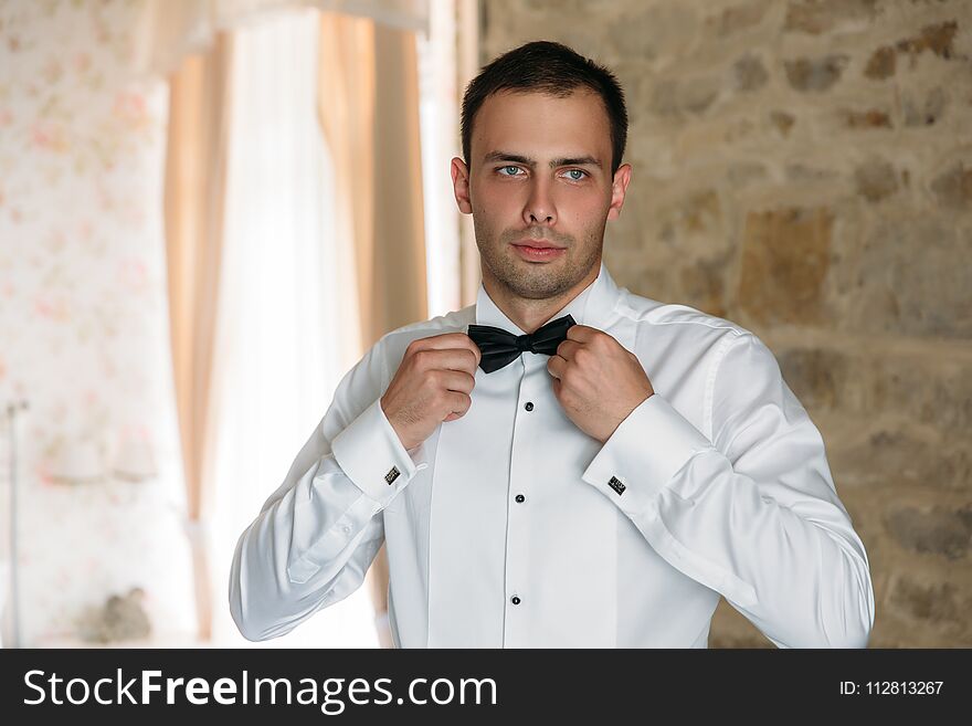 Morning of the groom. Businessman morning preparation. Young and handsome groom getting dressed in a wedding shirt holding bow-tie.