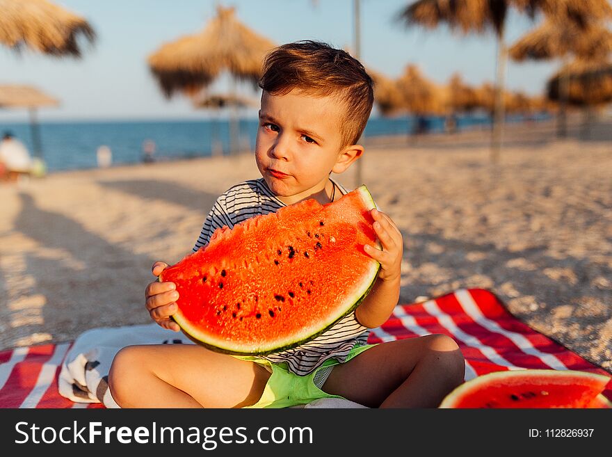 Funny little boy eating watermelon on the beach.Huge piece of watermelon it`s hard to carry it.