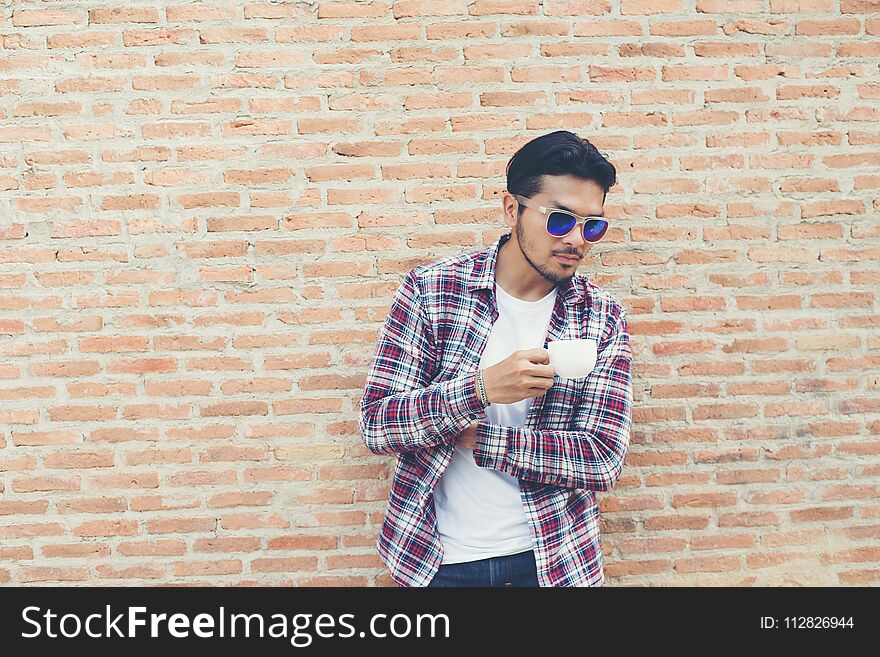 Handsome hipster young man holding coffee cup and looking away while standing at the street against brick wall background.