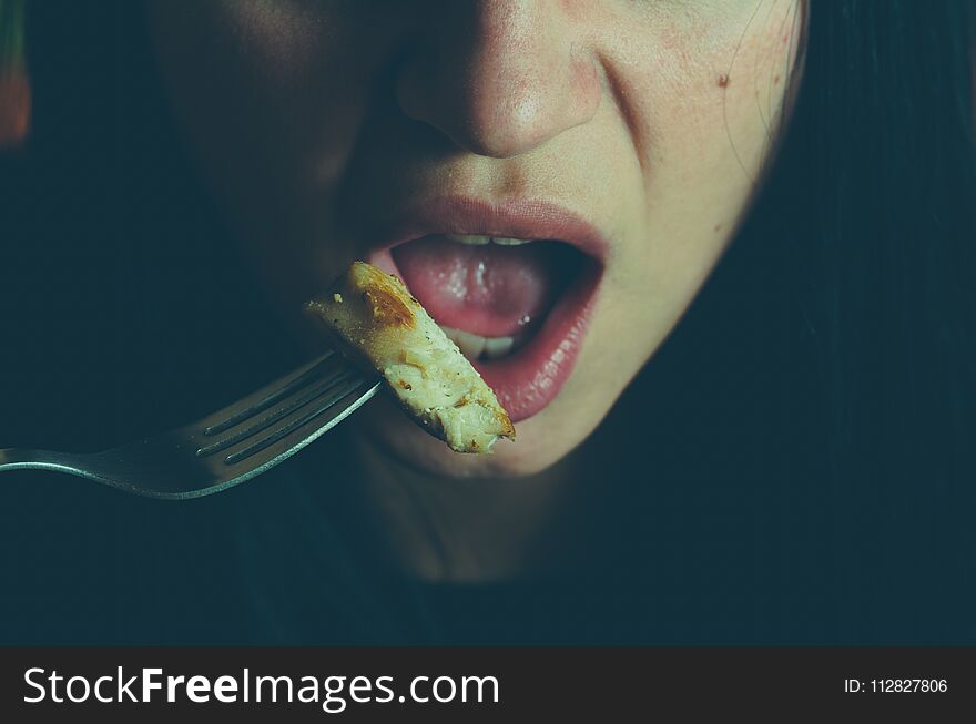 Part of the girls face with mouth and tongue holding fork with chicken meat eating in the night, hiding from other people, illuminated with light from the fridge, obesity concept, selective focus.