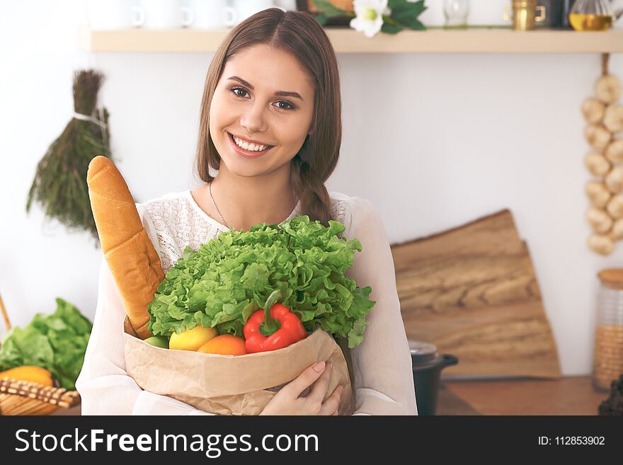 Young happy woman holding paper bag full of vegetables and fruits while smiling. Girl have made shopping and ready for cooking in kitchen.