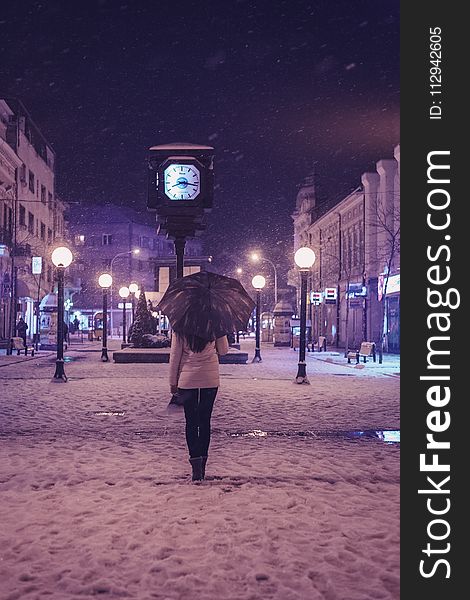 Person In White Long-sleeved Top And Black Pants Under Black Umbrella Standing Near Street Clock