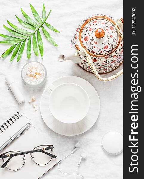 Flat lay cozy morning inspiration planning female table - teapot, tea cup, notepad, glasses, lipstick, face cream, green leaf flow