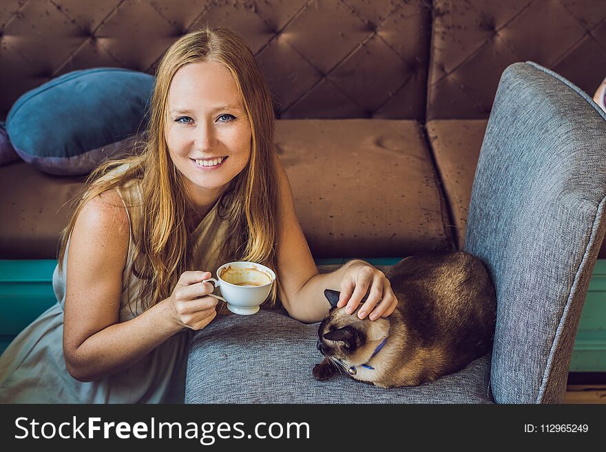 Young woman is drinking coffee and stroking the cat. Against the backdrop of the sofa scratched by cats.