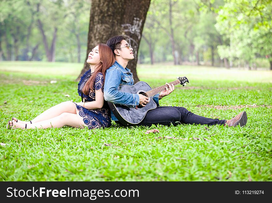 Asian couple in love playing acoustic song guitar sitting on grass in the park