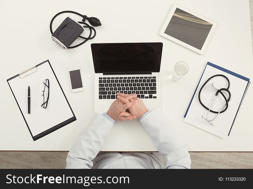 Doctor sitting at office desk with medical equipment and typing on his laptop with blank screen, top view, copy space. Doctor sitting at office desk with medical equipment and typing on his laptop with blank screen, top view, copy space