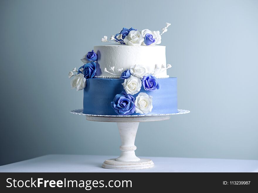 Beautiful two tiered white and blue wedding cake decorated with flowers sugar roses on the table. The concept of elegant holiday desserts