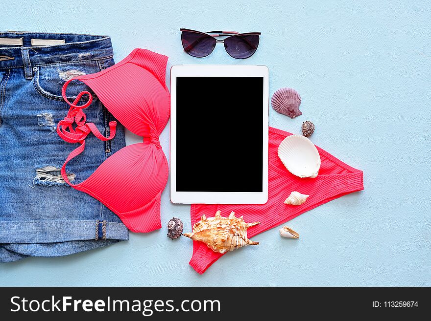 Female summer bikini swimsuit and accessories on blue with tablet and sunglasses.