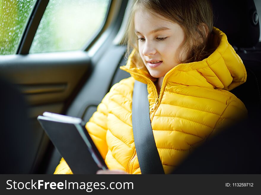 Adorable girl sitting in a car and reading her ebook on rainy autumn day. Child entertaining herserf on a road trip. Traveling by car with kids.