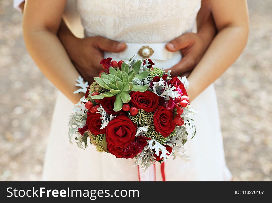 The bridegroom embraces the bride`s waist, a wedding bouquet in her hands, in focus a bouquet. the bridegroom embraces the bride and the wedding bouquet